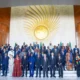 A wide-angle portrait of African Heads of State and international dignitaries, including UN Secretary-General António Guterres and Italian Prime Minister Giorgia Meloni, posing for the official family photo at the 39th African Union Summit in Addis Ababa to discuss African Union strengths and weaknesses. The leaders are gathered in the lobby of the Nelson Mandela Plenary Hall in front of the large golden AU emblem wall.